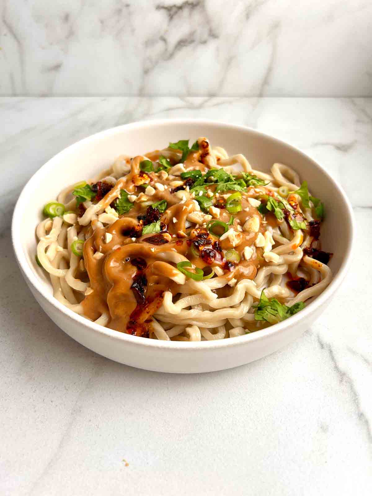 bowl of noodles with peanut sauce, cilantro, green onions, chili oil, and chopped peanuts sprinkled on top.