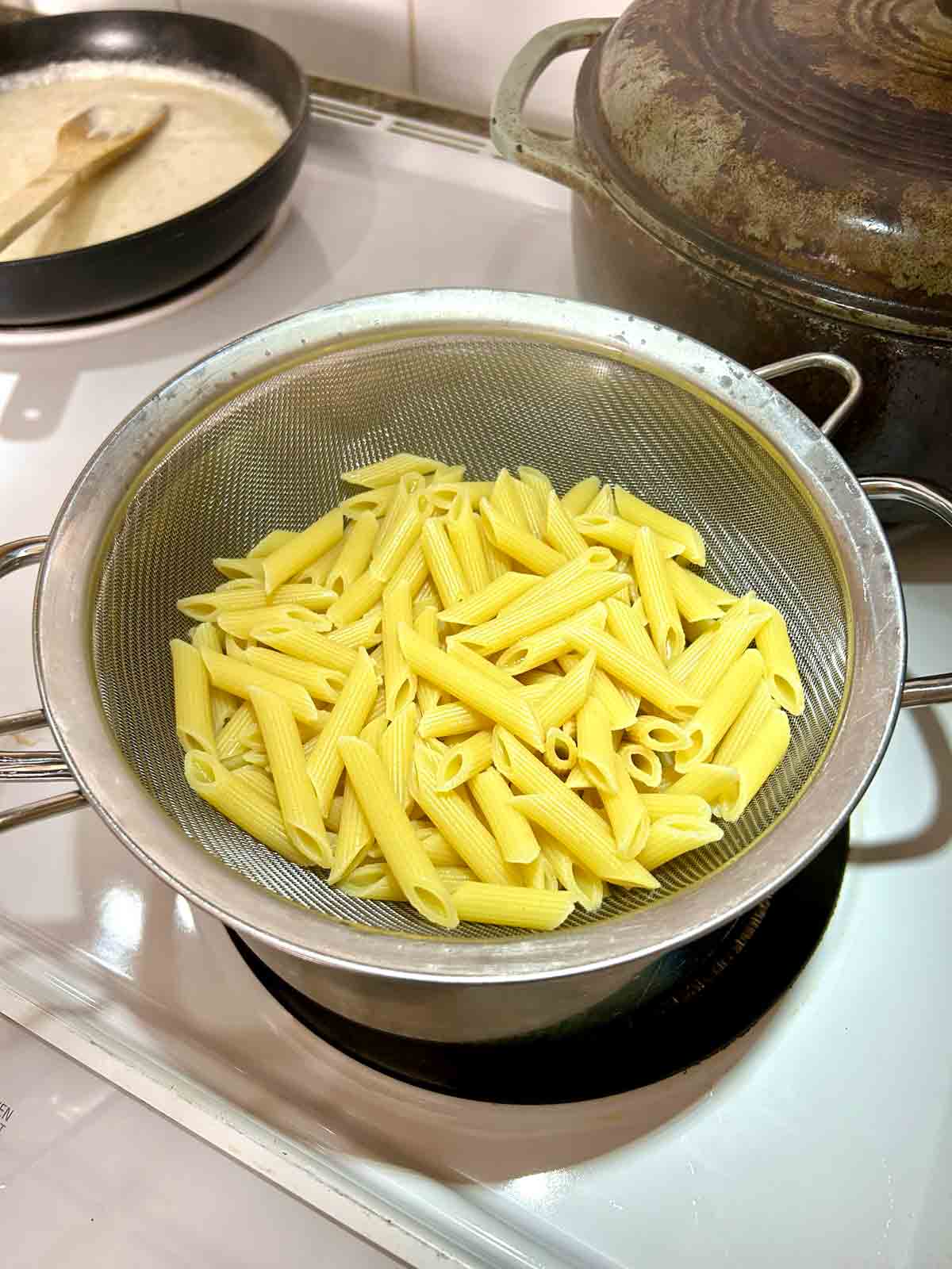 cooked penne pasta draining in strainer over pot.