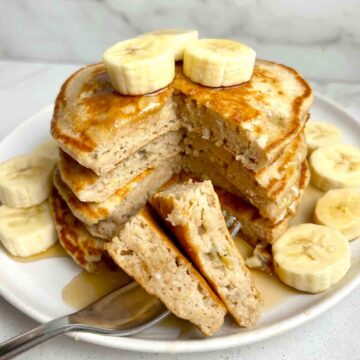 stack of banana pancakes cut through the middle to show the inside; fork piercing two pieces and fresh bananas added on plate.