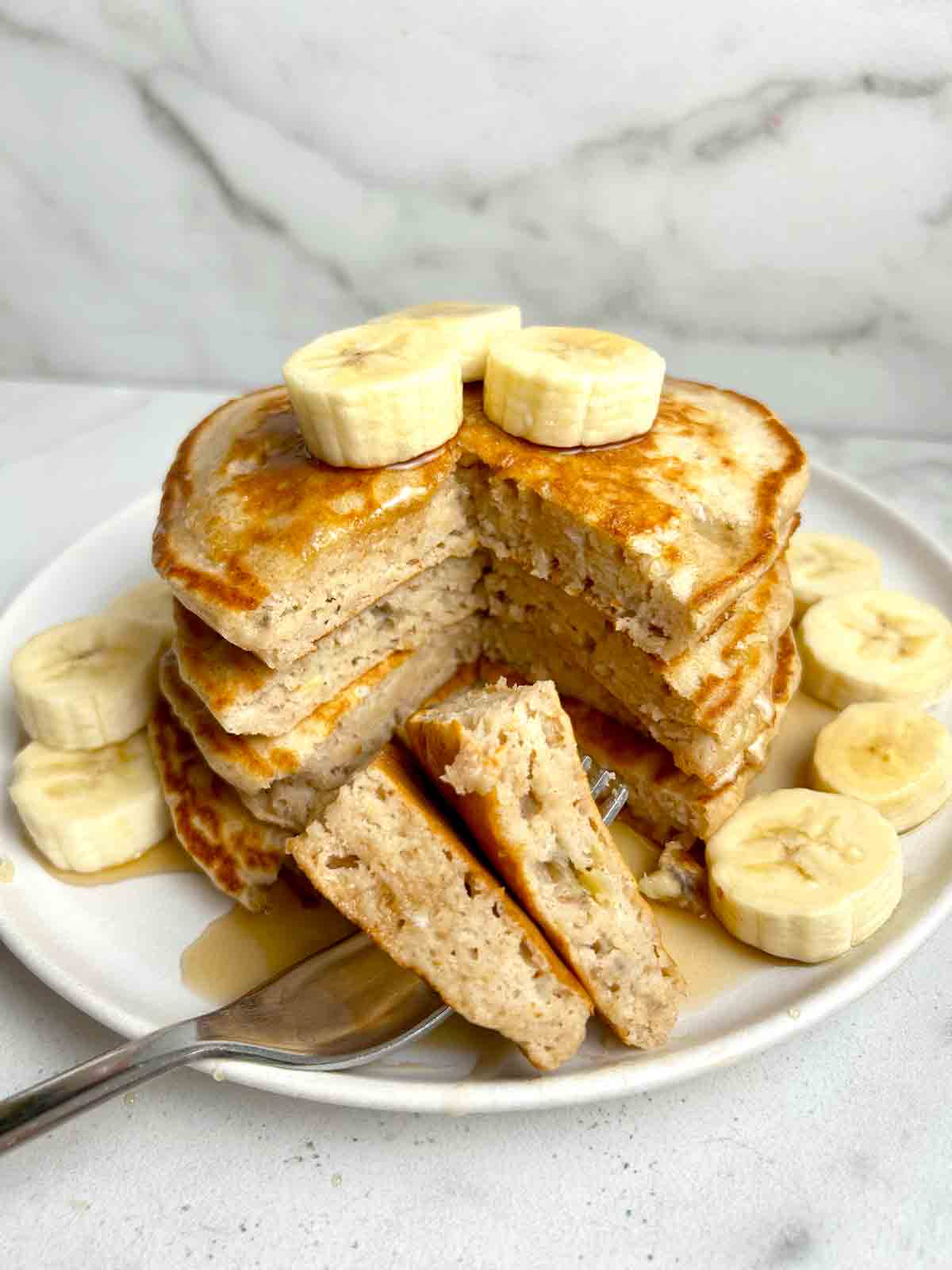 stack of banana pancakes cut through the middle to show the inside; fork piercing two pieces and fresh bananas added on plate.
