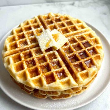 close up of 3 waffles stacked together; maple syrup, powdered sugar, and pat of butter on top.