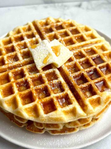 close up of 3 waffles stacked together; maple syrup, powdered sugar, and pat of butter on top.