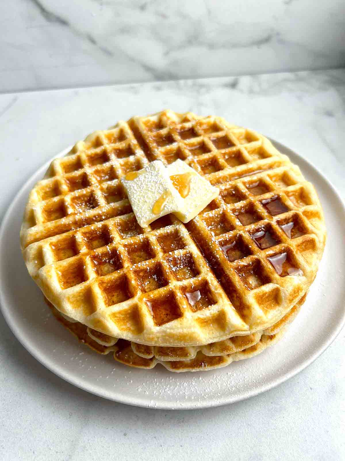 close up of 3 waffles stacked together; maple syrup, powdered sugar, and pat of butter on top.