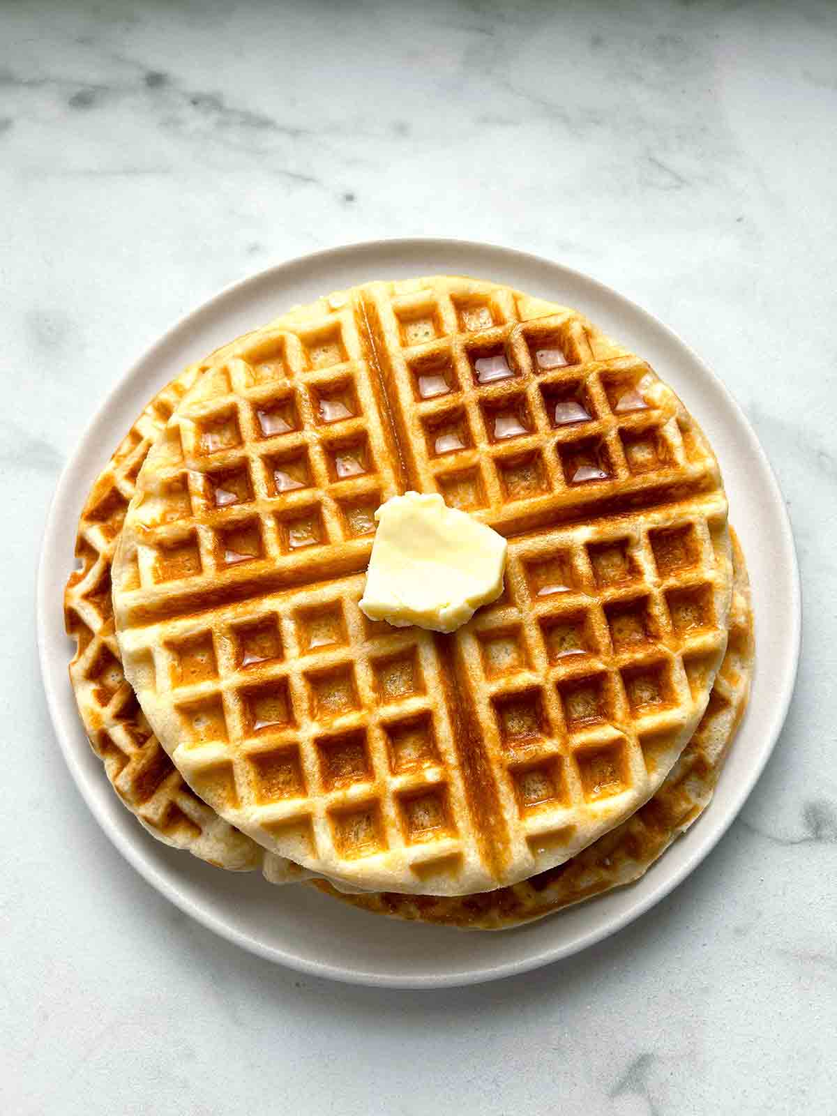 waffles stacked together on plate; pat of butter and maple syrup on top.