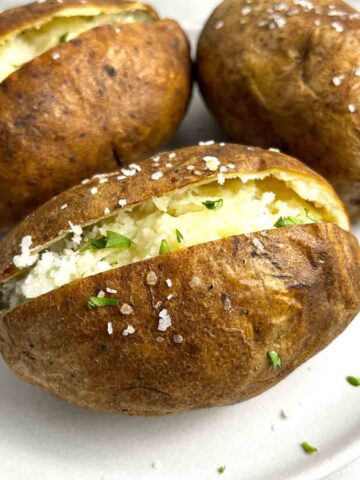 close up of plate of baked potatoes with one with slit cut through top, garnished with parsley and butter.