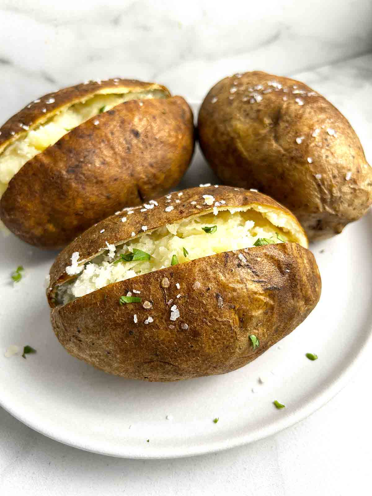 close up of plate of baked potatoes with one with slit cut through top, garnished with parsley and butter.