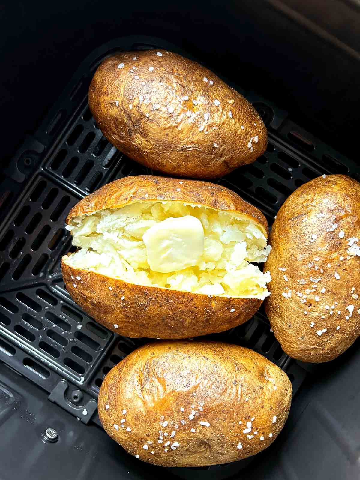 close up of baked potatoes in air fryer basket; one is split open with slab of butter on top.