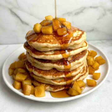 syrup being poured on stack of apple pancakes with diced apples around plate.