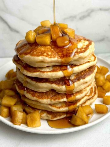 syrup being poured on stack of apple pancakes with diced apples around plate.