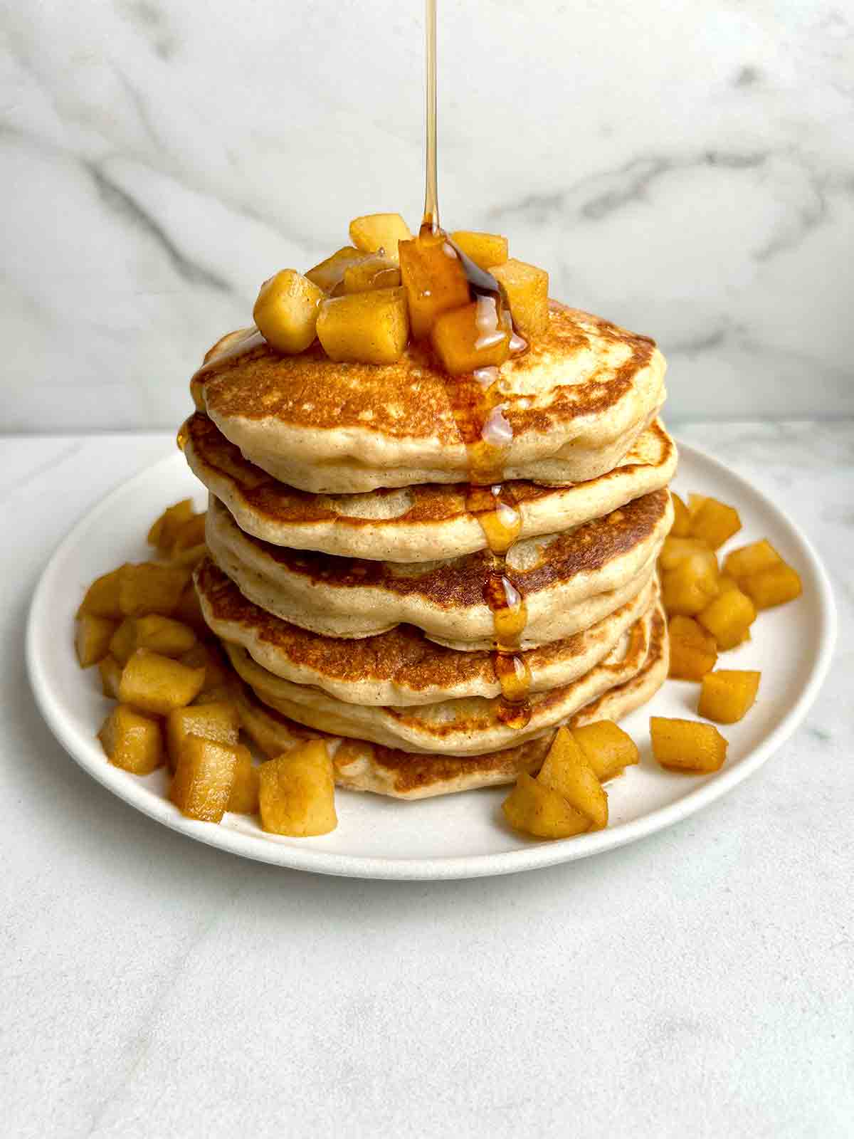 syrup being poured on stack of apple pancakes with diced apples around plate.