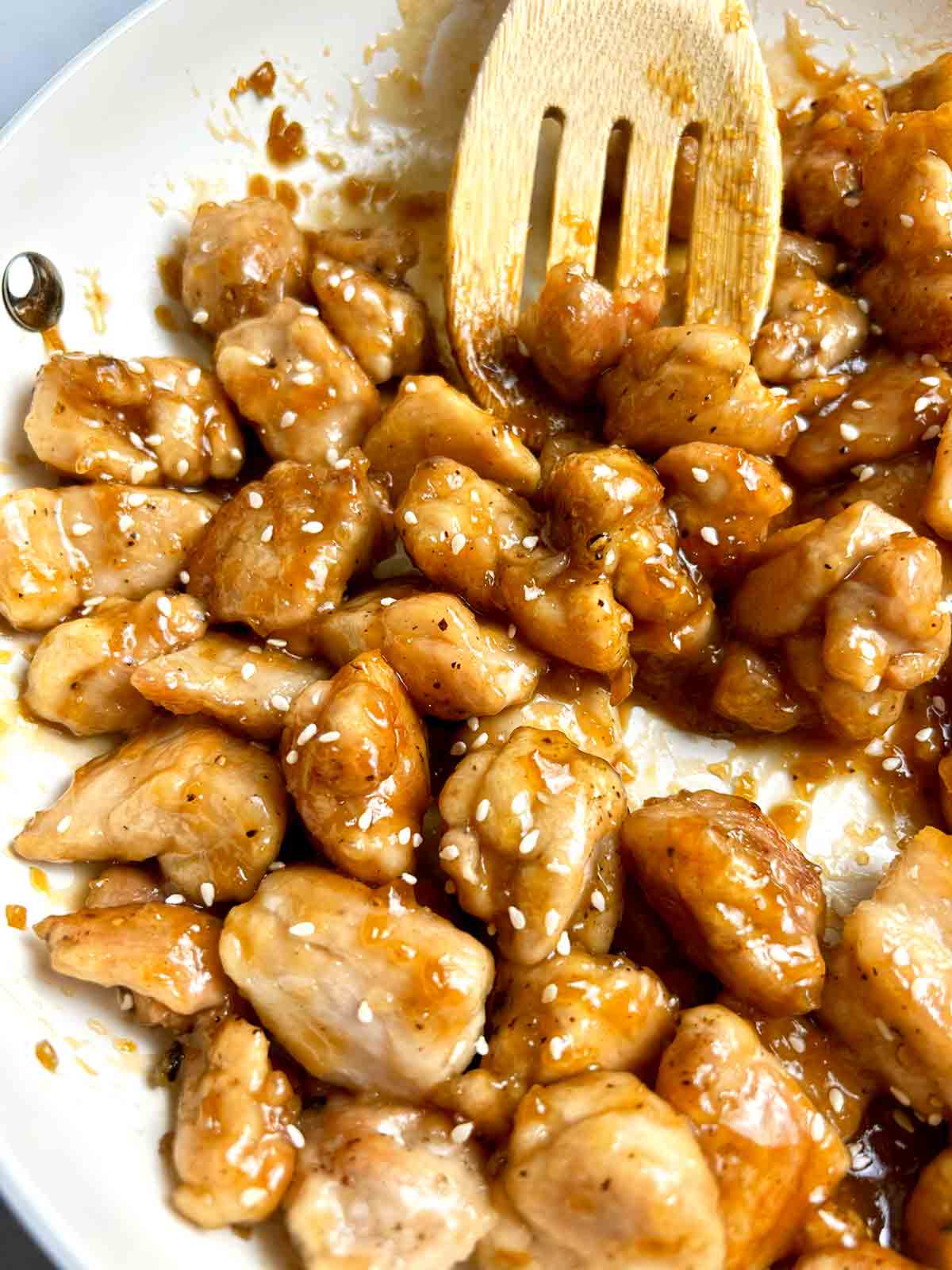 close up of honey garlic chicken bites in skillet; wooden spatula lifting a few pieces from pan.